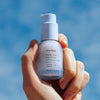 Close up of hand holding Ursa Major Sunny Start Eye Serum (0.5 oz) against a blue sky background