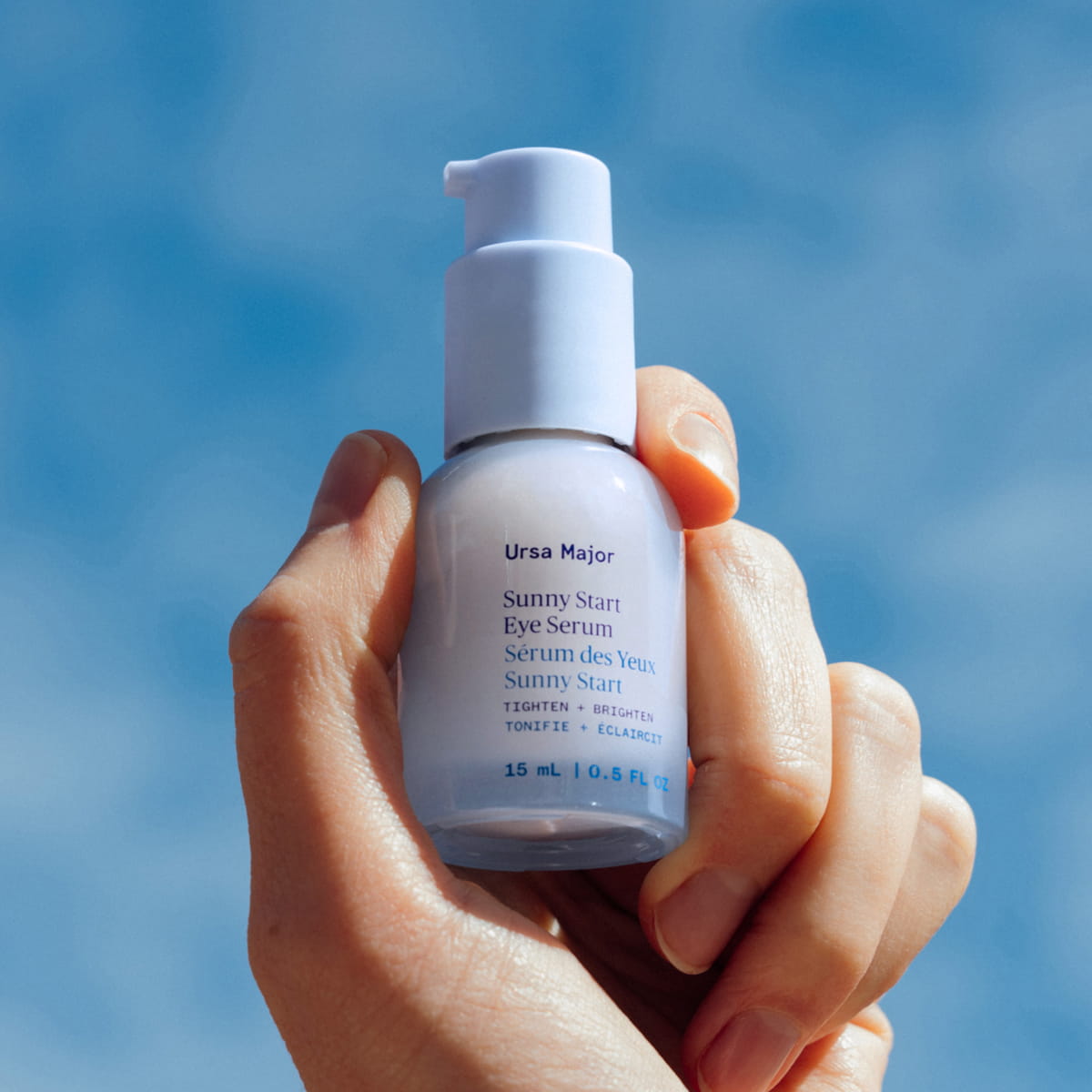 Close up of hand holding Ursa Major Sunny Start Eye Serum (0.5 oz) against a blue sky background