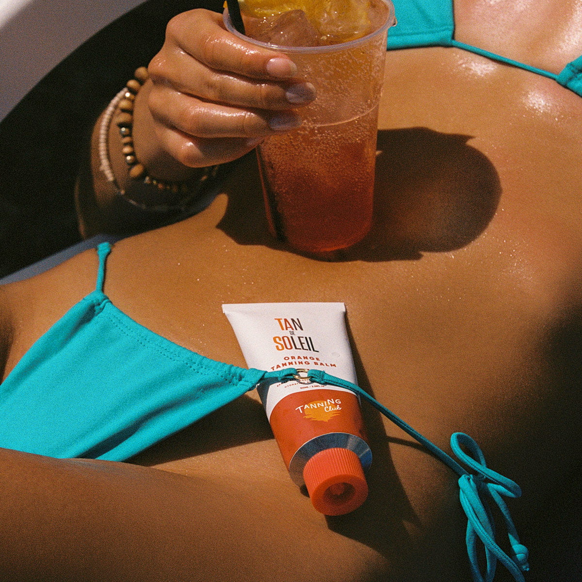 Woman in bikini with Tanning Club Tan de Soleil Orange Tanning Balm SPF 6 (3.38 oz) under strap of bikini bottom holding an iced drink