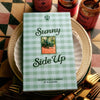 Flamingo Estate Sunny Side Up Recipe Book (1 pc) - styled on a stack of plates with gold cutlery and jars in the background.