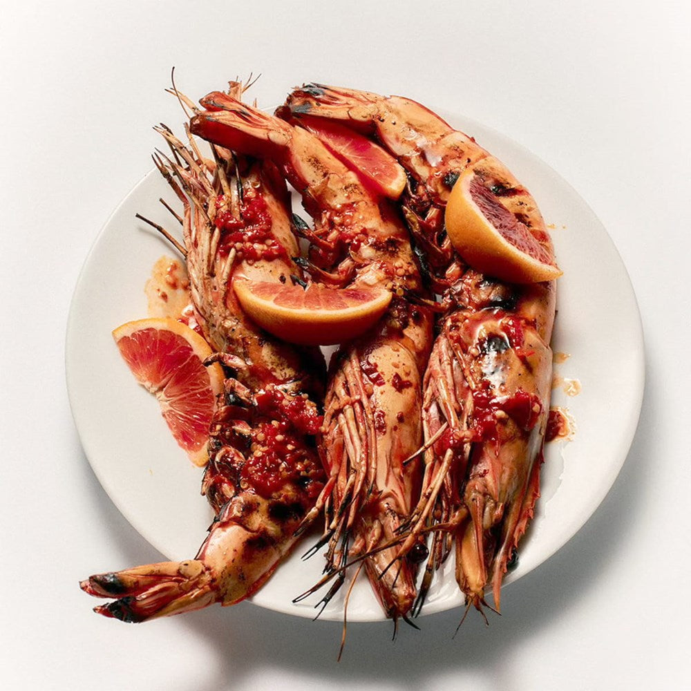 Flamingo Estate Sunny Side Up Recipe Book (1 pc) - Grilled shrimp cocktail garnished with red grapefruit slices and a red sauce, served on a white plate against a light gray background.
