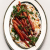 Flamingo Estate Sunny Side Up Recipe Book (1 pc) - A top-down view of a prepared food dish featuring roasted carrots served over a white creamy base, garnished with chopped green herbs, walnuts, and a drizzle of sauce on an oval white plate with a gold rim.