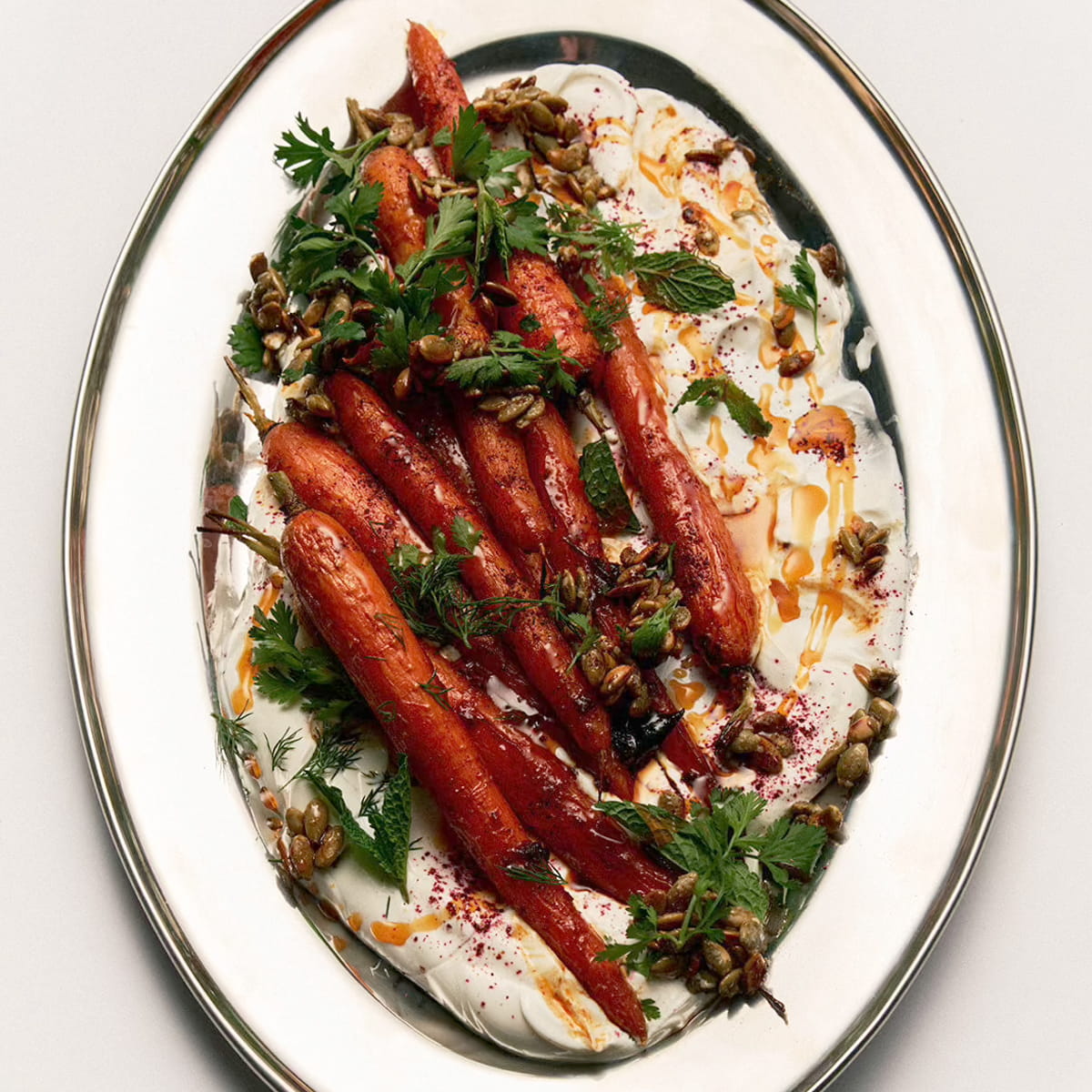 Flamingo Estate Sunny Side Up Recipe Book (1 pc) - A top-down view of a prepared food dish featuring roasted carrots served over a white creamy base, garnished with chopped green herbs, walnuts, and a drizzle of sauce on an oval white plate with a gold rim.