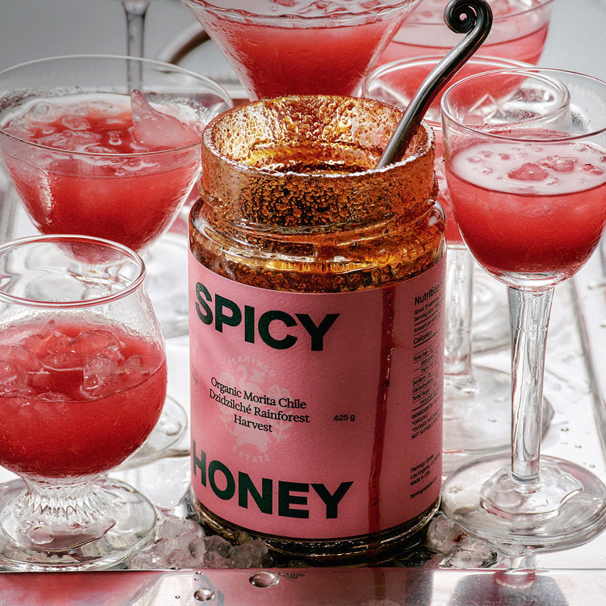 Flamingo Estate Sunny Side Up Recipe Book (1 pc) - A jar of 'Spicy Honey' with a pink label and a gold glittery lid sits on a silver tray surrounded by glasses of a pink-red beverage.