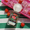 Lifestyle shot of Maison Matine Bain De Minuit Eau de Parfum (100 ml) bottle, placed on a green and white striped fabric next to red cherries and a pink ice cube tray with cherries inside.