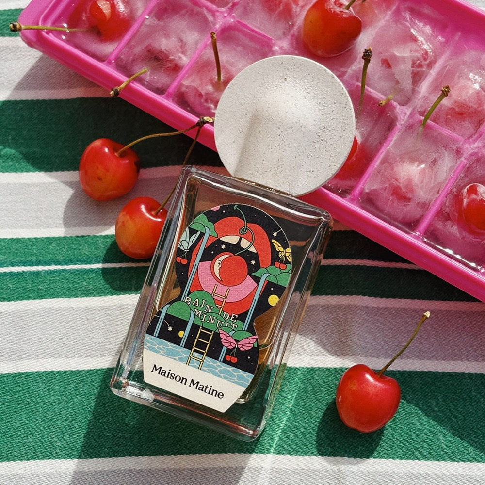 Lifestyle shot of Maison Matine Bain De Minuit Eau de Parfum (100 ml) bottle, placed on a green and white striped fabric next to red cherries and a pink ice cube tray with cherries inside.