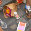 Maison Matine Shifumi Eau de Parfum (100 ml) and box on stone pavement featuring a brown paper flower bouquet, multiple shoes (checkered, leopard print), with wine glasses held by partially visible hands and legs.