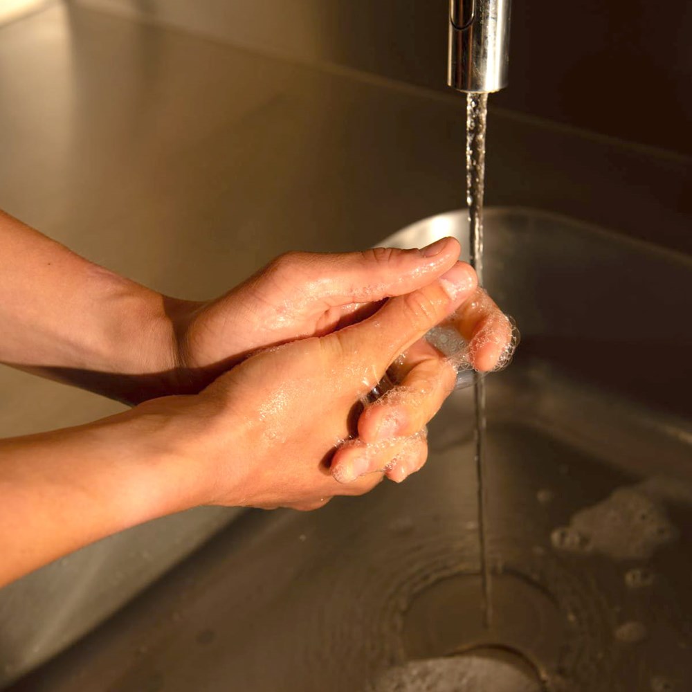 Naomi Los Angeles Terroir Hand Wash - close-up of a person washing hands under running water from a chrome faucet in a stainless steel sink.