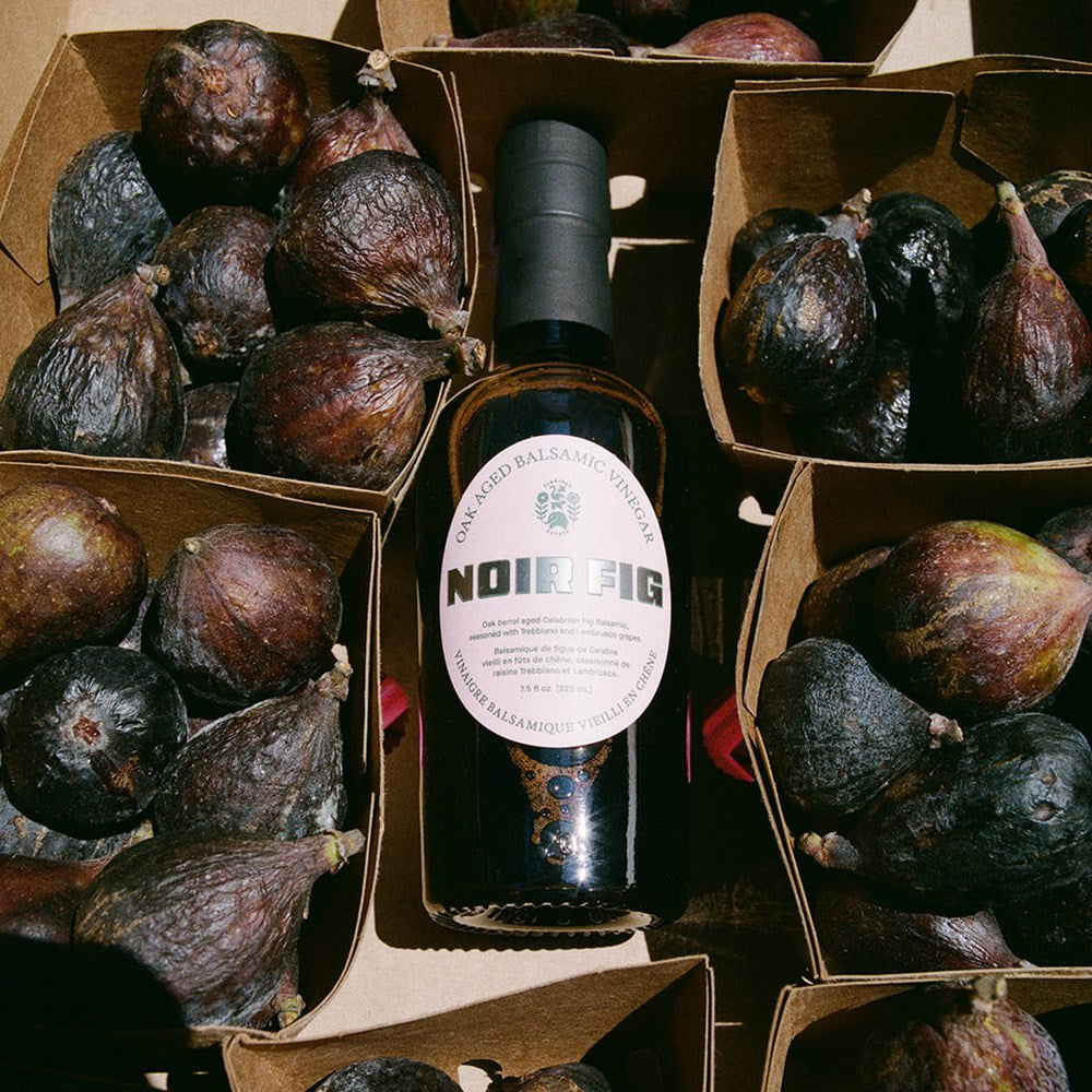Bottle of Flamingo Estate Noir Fig Balsamic Vinegar (222 ml) centered between multiple cardboard boxes filled with dark figs.