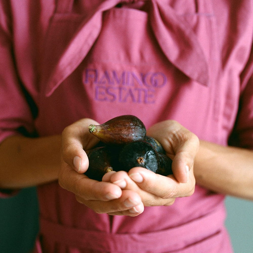 A person wearing a pink apron with 'FLAMINGO ESTATE' embroidered on the chest holds several dark figs in their hands.
