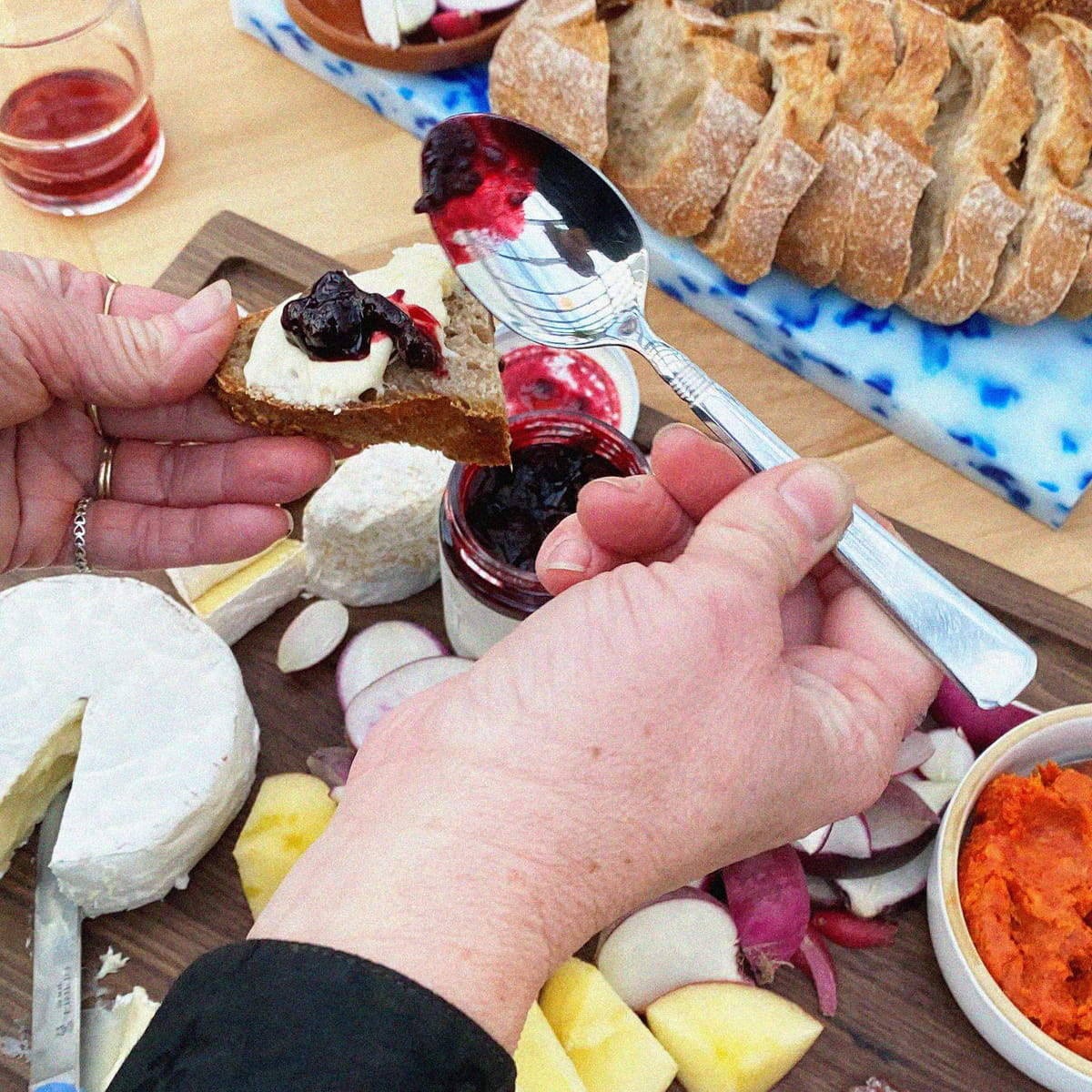 A close-up lifestyle shot of a charcuterie board being prepared, with hands spreading C. Cassis Salted Currant Compote onto a piece of bread, surrounded by sliced bread, cheese, radishes, and fruit on a wooden board.
