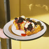 A hand holds a white plate with a black rim with two golden-brown pastries with C. Cassis Salted Currant Compote on top set against a yellow wall with strong sunlight and shadows.
