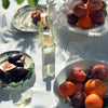 Lifestyle shot of Parfums de Nicolai Fig Tea Intense (100 ml) stands on a white tablecloth surrounded by fruit plates, glassware, and natural sunlight shadows.