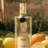 Lifestyle shot of Parfums de Nicolai Fig Tea Intense (100 ml) centered and surrounded by fresh lemons and oranges against a rustic stone wall and green foliage background.