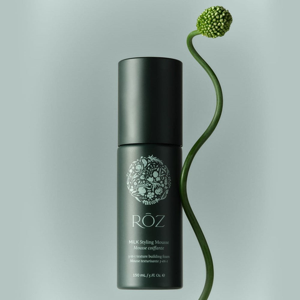 Lifestyle shot of Roz Milk Styling Mousse (150 ml) with a green plant stem and seed pod against a light green background.