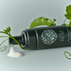 Lifestyle shot of Roz Milk Styling Mousse (150 ml) lies on its side with mousse dripping from the nozzle. Green leaves and vines are artistically arranged around the bottle against a light green background.