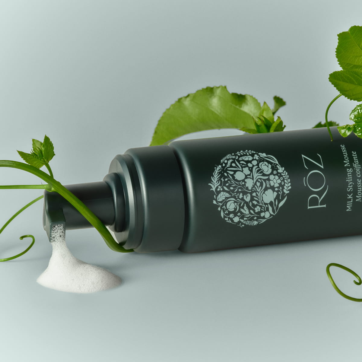 Lifestyle shot of Roz Milk Styling Mousse (150 ml) lies on its side with mousse dripping from the nozzle. Green leaves and vines are artistically arranged around the bottle against a light green background.