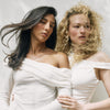 Two women posing in matching off-the-shoulder white long-sleeve tops against a light neutral studio background. Both with Roz Milk Styling Mousse used to style their hair