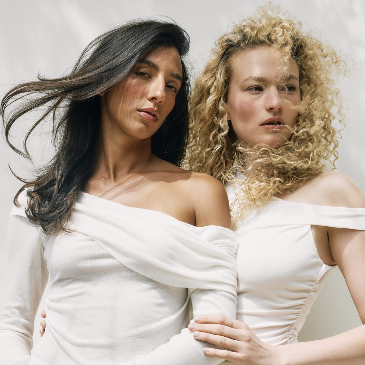 Two women posing in matching off-the-shoulder white long-sleeve tops against a light neutral studio background. Both with Roz Milk Styling Mousse used to style their hair