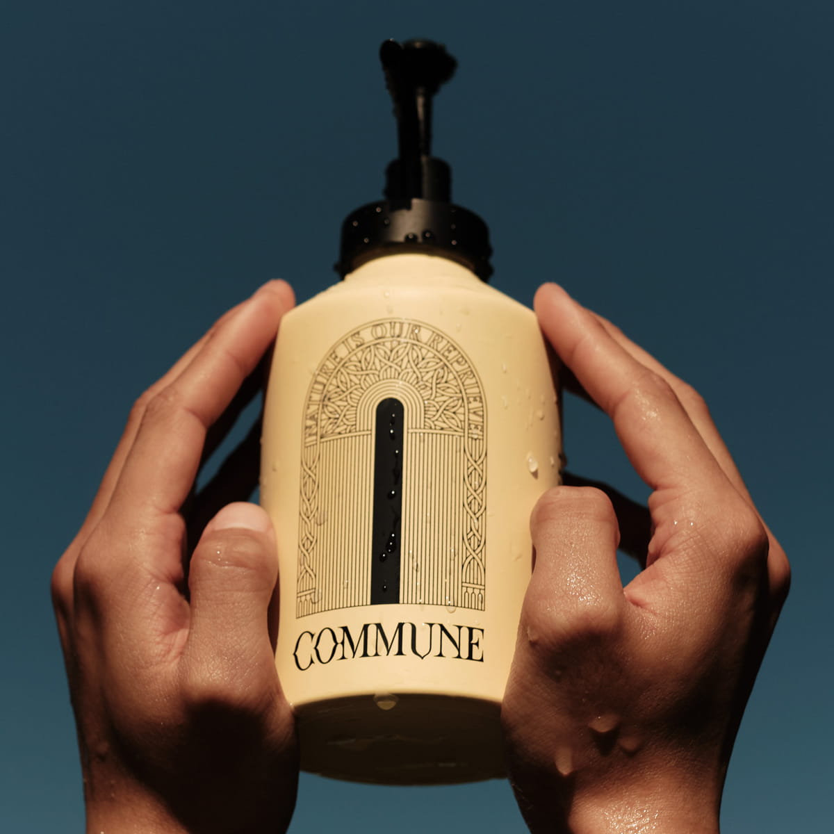 A person's hands holding Commune Seymour Hand Cream & Reusable Pump (500 ml) against a dark blue sky
