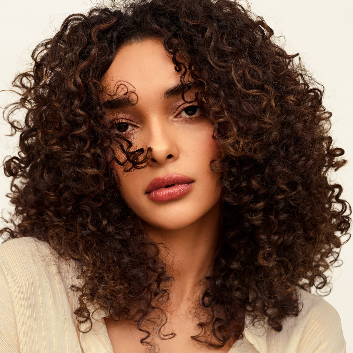 A close-up portrait of a woman with Innersense Organic Beauty Overnight Repair Serum used, showing voluminous, tightly curled dark brown hair
