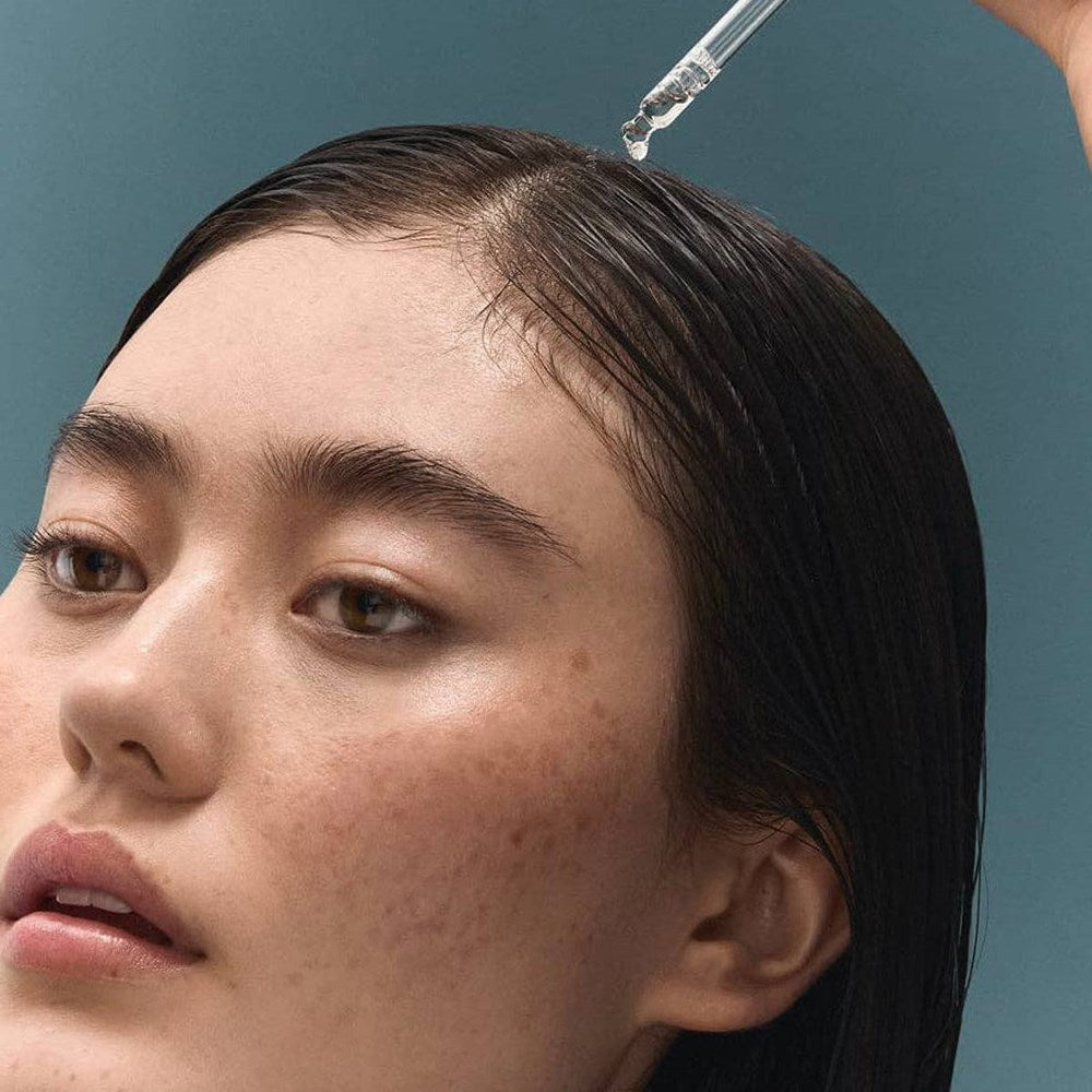 Close-up of a woman's face with wet dark hair receiving a dropper application of BASE-K The Bamboo Tonic Hair Density Serum