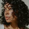 A close-up of a woman with voluminous, dark, curly hair using 14th Night Natural Surf Spray
