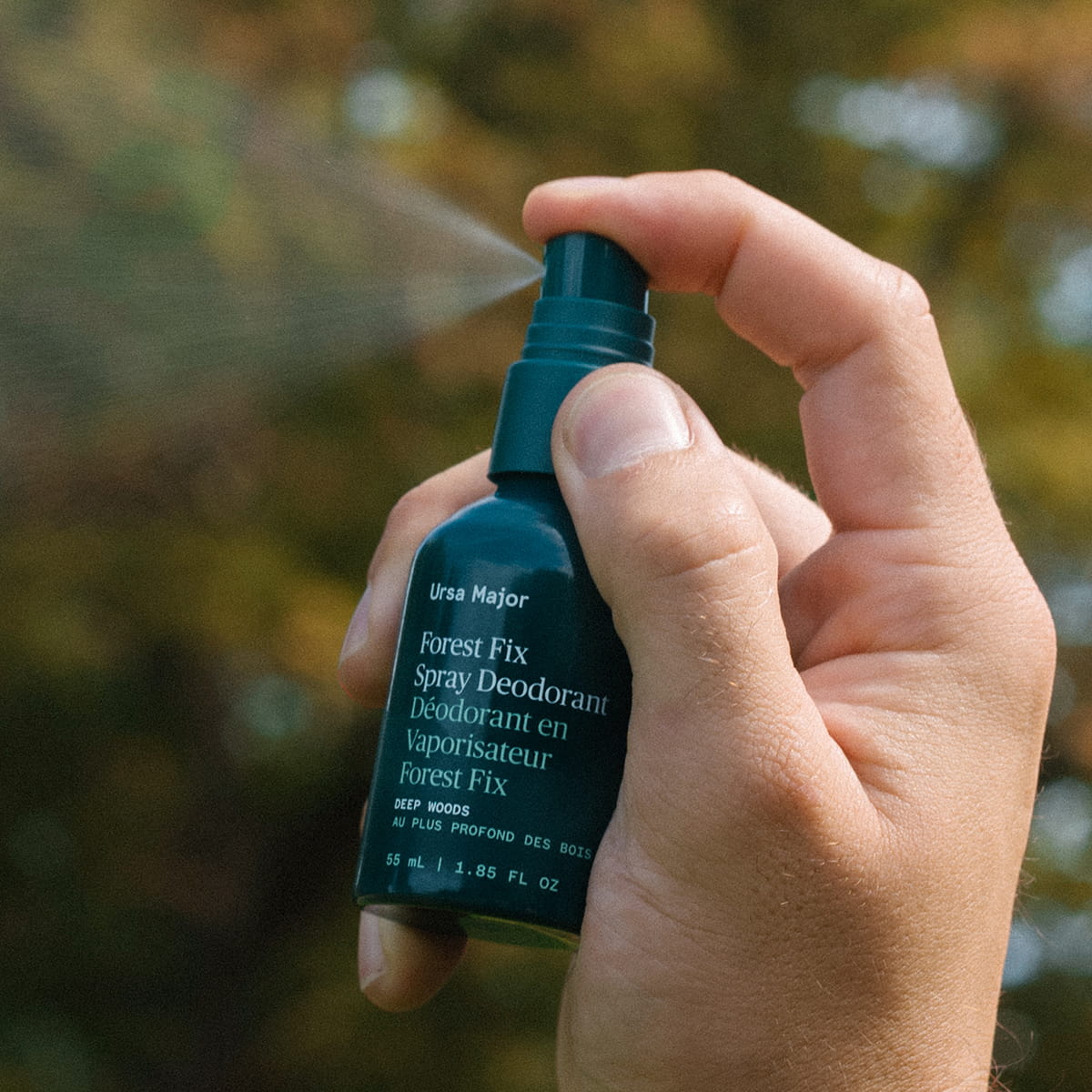 A hand holding Ursa Major Forest Fix Spray Deodorant (1.85 oz) with the spray mist visible, against a blurred outdoor background.