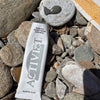 Activist Manuka Honey Glacial Clay Mask (80 ml) lies diagonally on a bed of smooth river stones outdoors. The cap is off and the mask swatch is on top a flat rock