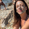 A woman on a rocky beach with water in the background, wearing a layer of Activist Manuka Honey Glacial Clay Mask on her face
