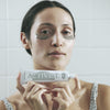 A woman holds Activist Manuka Honey Glacial Clay Mask (80 ml) in front of her face, with mask applied under her eyes
