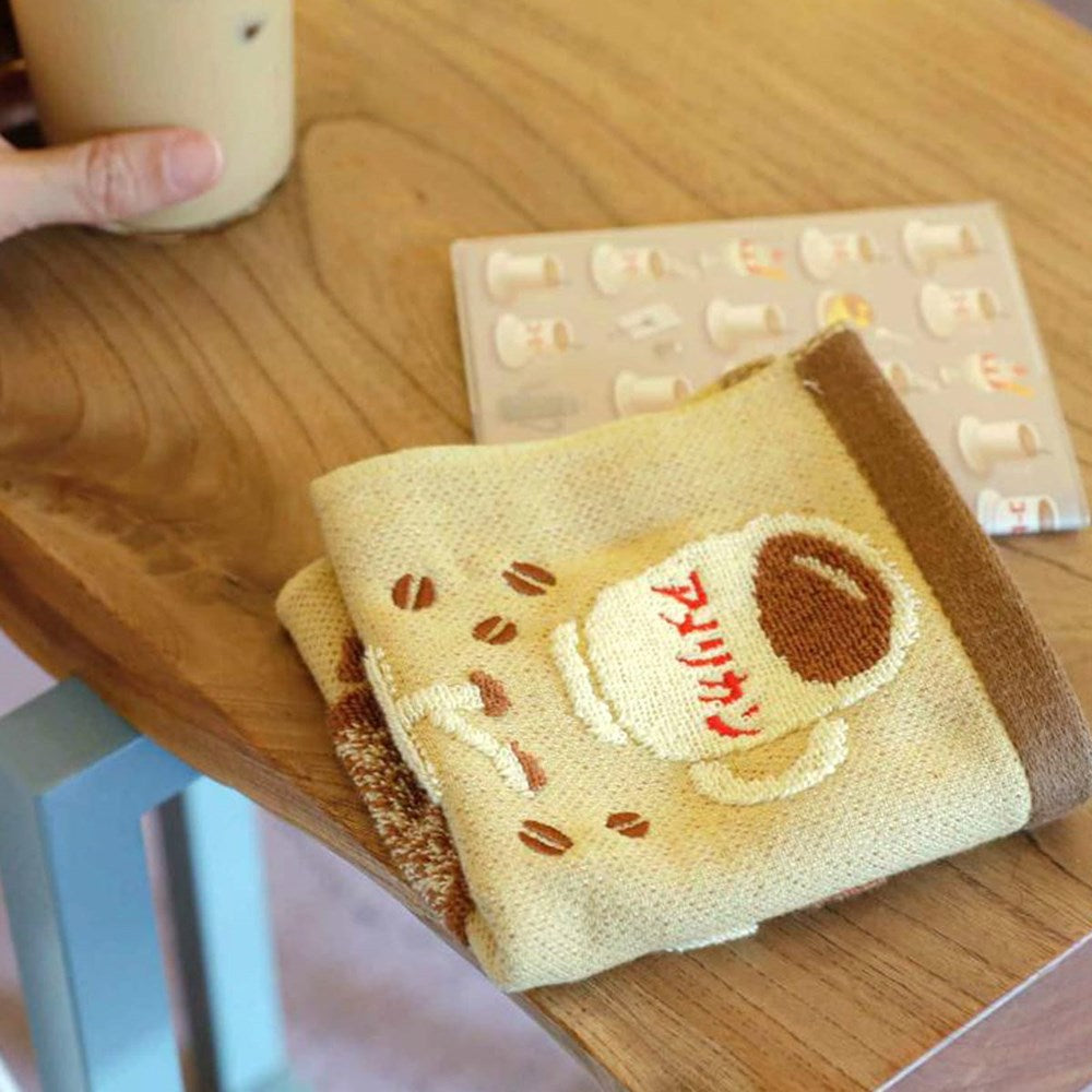 A close-up of a folded Hightide Hand Towel - Coffee (1 pc), placed on a wooden table next to a hand holding a cup