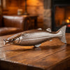 Double Neat Whiskey Trout Flask (1 pc)  resting on a rustic wooden table in a warm, cozy indoor setting with a fireplace in the background.