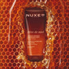 A tube of Nuxe Reve de Miel Deliciously Nourishing Body Scrub (200 ml) stands centered against a honeycomb and honey-themed background the scene is framed by dripping honey and golden-orange tones