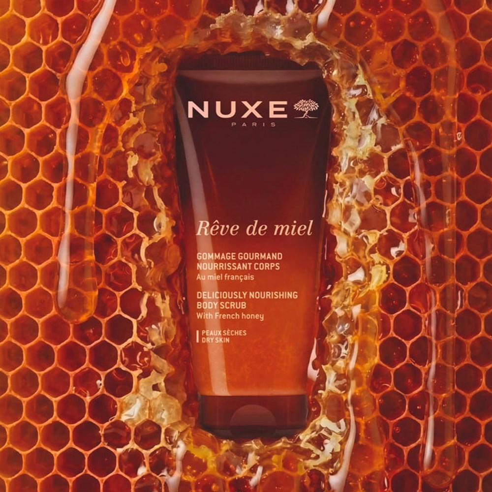 A tube of Nuxe Reve de Miel Deliciously Nourishing Body Scrub (200 ml) stands centered against a honeycomb and honey-themed background the scene is framed by dripping honey and golden-orange tones