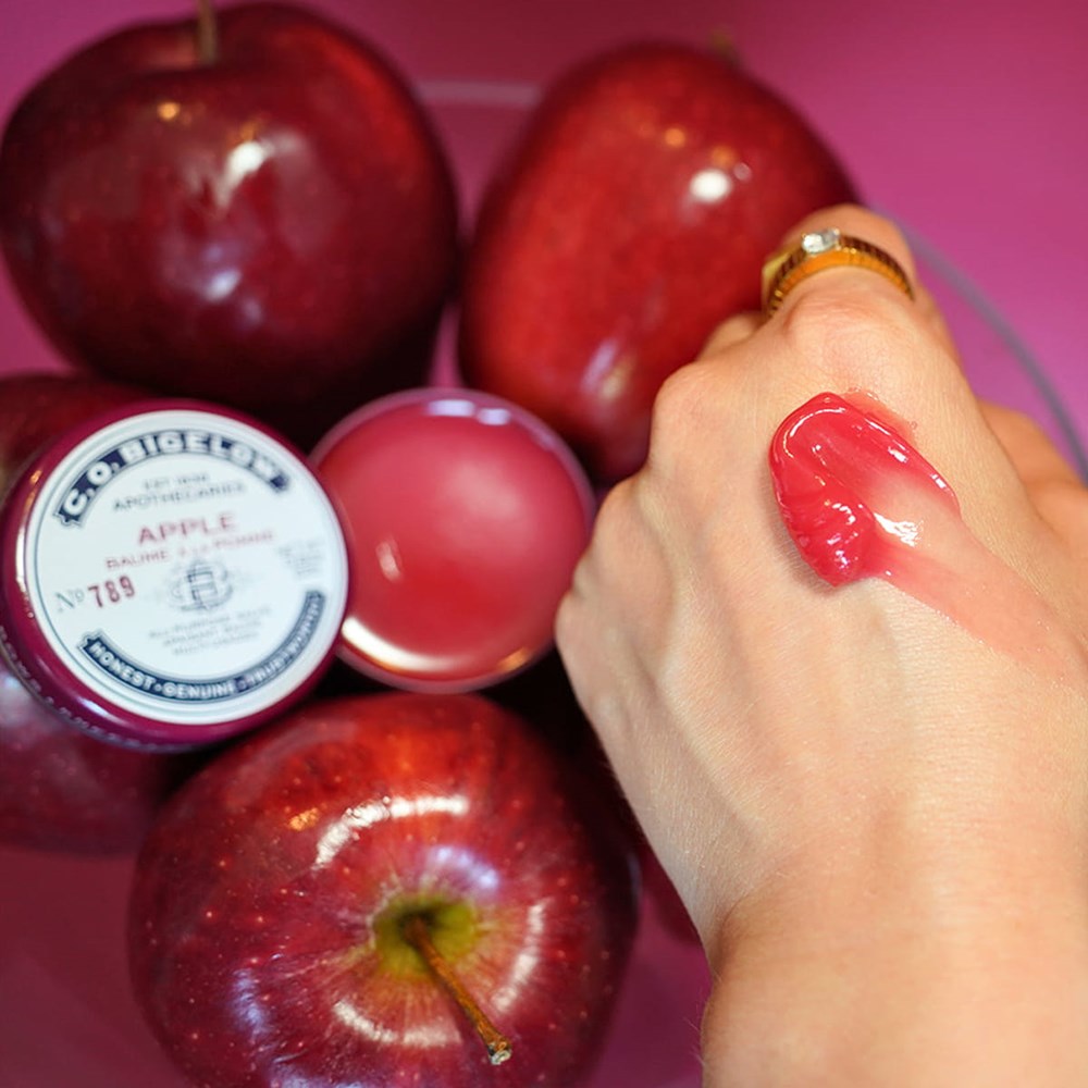 C.O. Bigelow Apple Salve Lip Tin (0.8 oz) - product shown as smear on hand next to opened product and red apples 