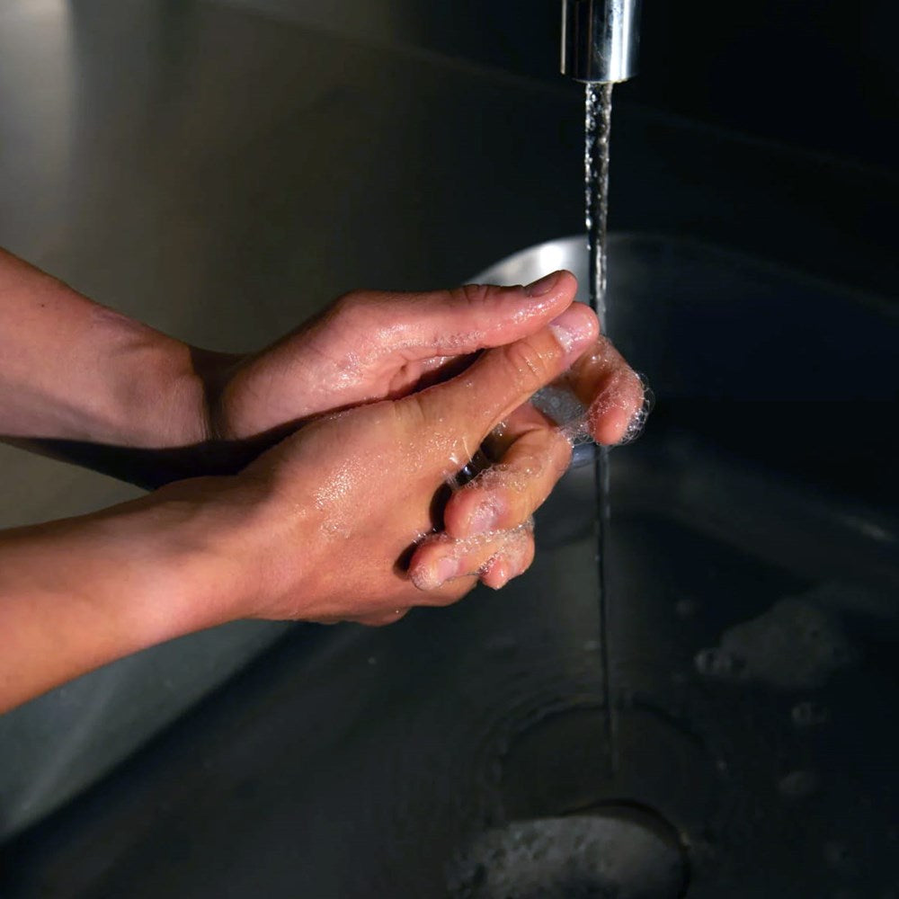 Close-up of a person's hands washing under a stream of water from a chrome faucet with Naomi After-Cooking Hand Scrub - Wood &amp; Yuzu
