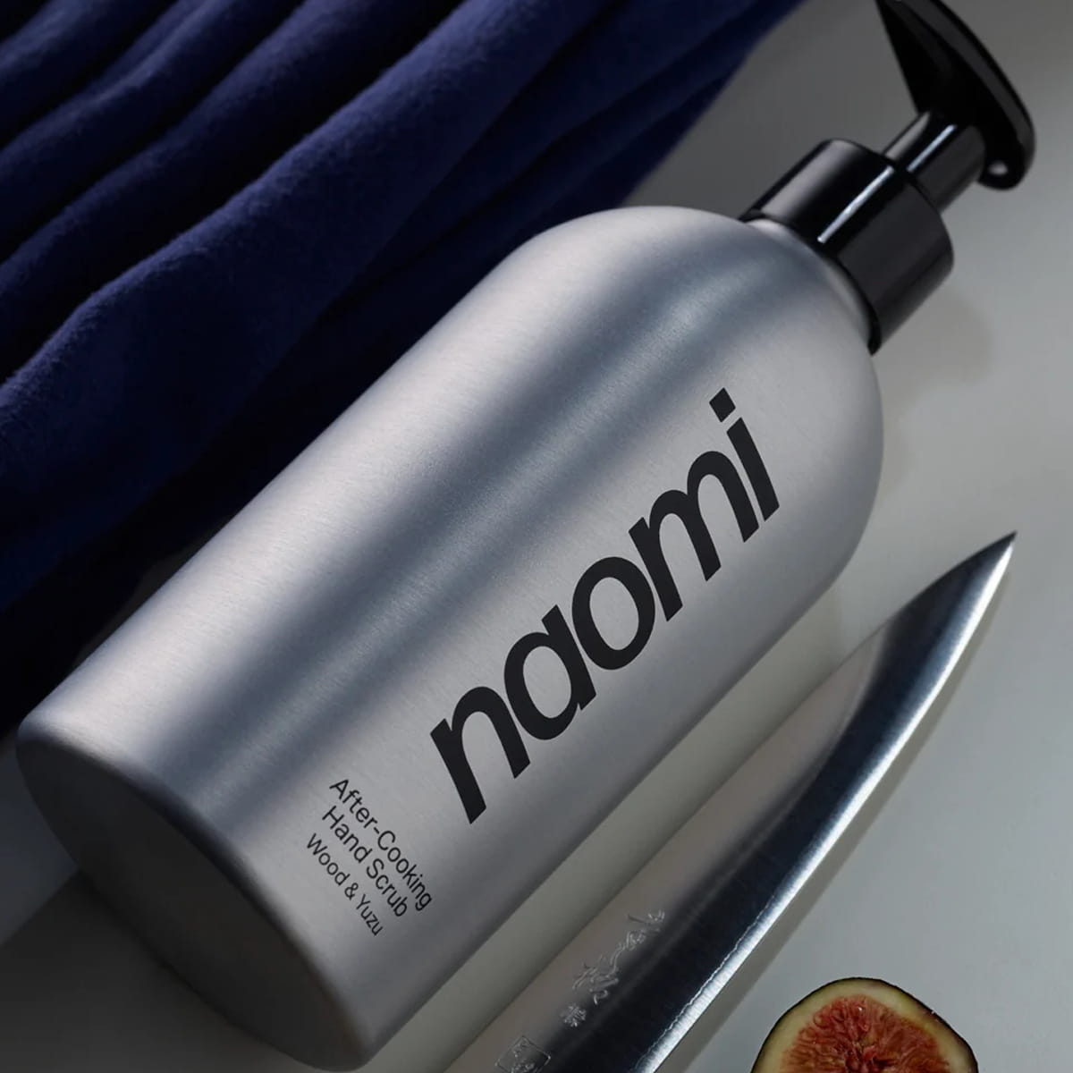 Naomi After-Cooking Hand Scrub - Wood &amp; Yuzu (500 ml) shown diagonally on a light surface with a dark blue textured cloth, next to a kitchen knife, and a fig slice