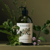 A bottle of Koala Eco Hand Wash - Rosalina & Peppermint (Glass Bottle) (16.9 oz), surrounded by botanical elements including green leaves, small purple flowers, and pine branches against a dark olive background.