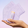 A hand holding VOESH New York Heel Moisturizing Heel Socks in purple and Solemate Heel Repair Balm in lavender against a soft pastel gradient background.