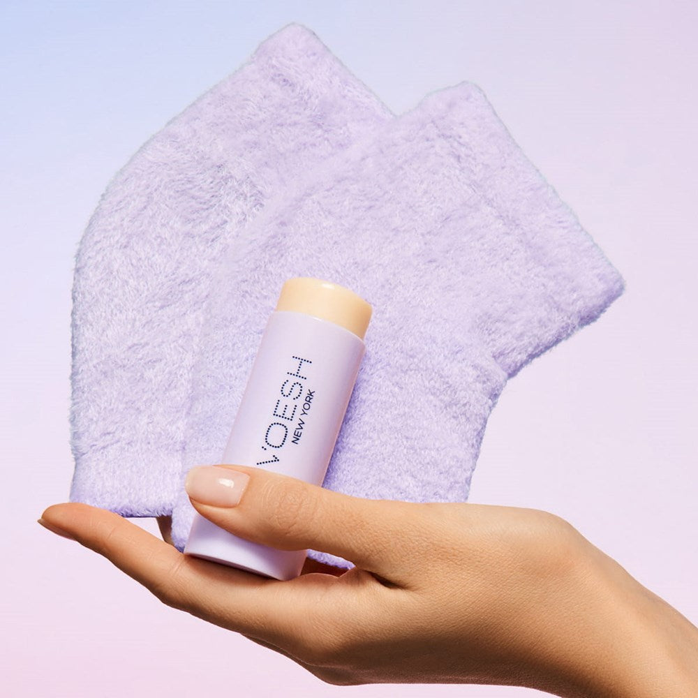 A hand holding VOESH New York Heel Moisturizing Heel Socks in purple and Solemate Heel Repair Balm in lavender against a soft pastel gradient background.