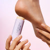 A close-up of a person holding a VOESH Solemate Heel Repair Balm in Lavender against their bare heel, with the other hand gently touching the heel.