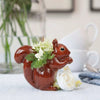 lifestyle shot of Quail Ceramics Squirrel Bud Vase (1 pc) shown side view with green foliage and white flowers in the vase and white flowers next to vase
