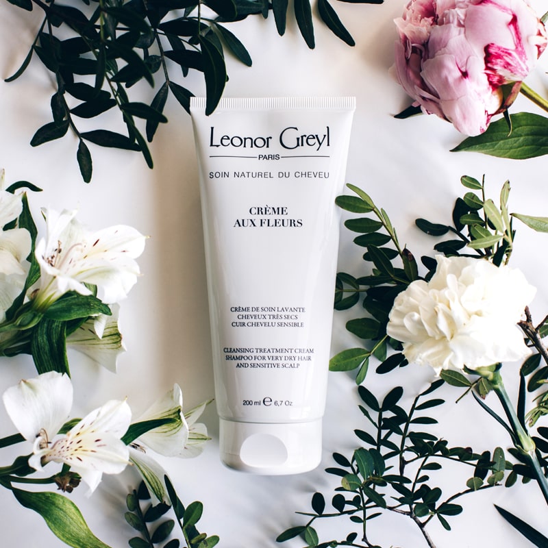 Lifestyle shot top view of Leonor Greyl Creme aux Fleurs Cream Washing Cream (200 ml) with white and pink flowers and leaves in the background