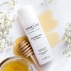 Lifestyle shot top view of Leonor Greyl Shampooing au Miel (120 ml) with honey and delicate white flowers in the background