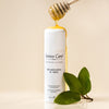 Lifestyle shot of Leonor Greyl Shampooing au Miel (120 ml) with honey being drizzled on top of bottle and leaves in the foreground