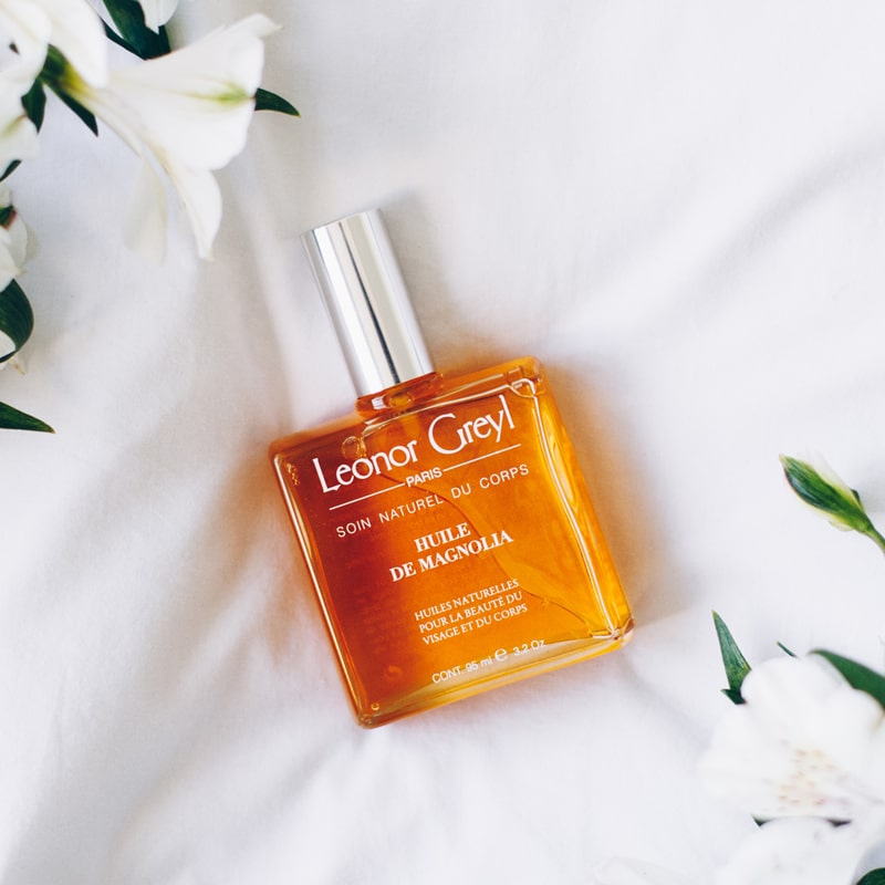 Lifestyle shot top view of Leonor Greyl Magnolia Beauty Oil (95 ml) with white flowers and leaves in the background