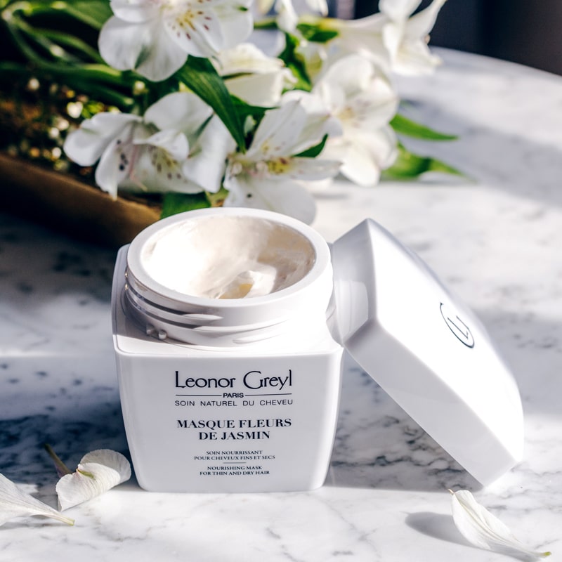 Lifestyle shot of Leonor Greyl Masque Fleurs de Jasmin Jasmine Conditioner (200 ml) with lid off showing color and texture of masque and white flowers and petals in the background and foreground