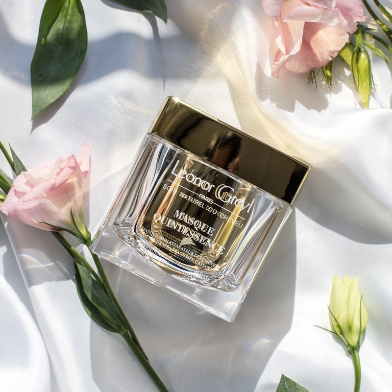Lifestyle shot top view of Leonor Greyl Masque Quintessence (200 ml) with pink and white flowers with leaves in the background