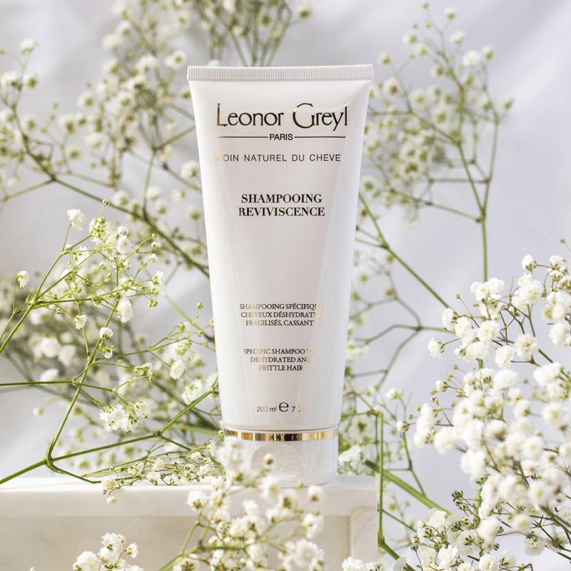 Lifestyle shot of Leonor Greyl Reviviscence Shampoo( 200 ml) with white flowers in the background